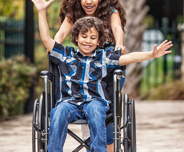 Boy smiles as his sister pushes him in his wheelchair outdoors