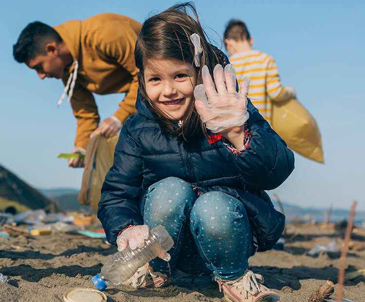 Young girl waving hello as she picks up litter from a beach.